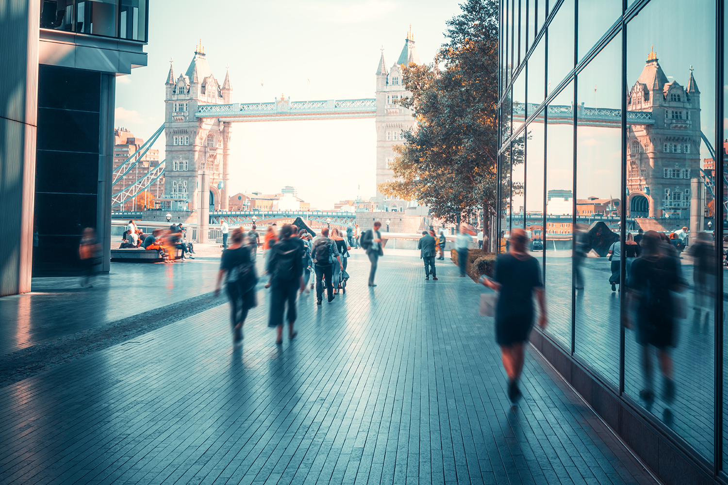 People walking through a modern commercial walkway between glazed buildings, with a landmark bridge in the distance.