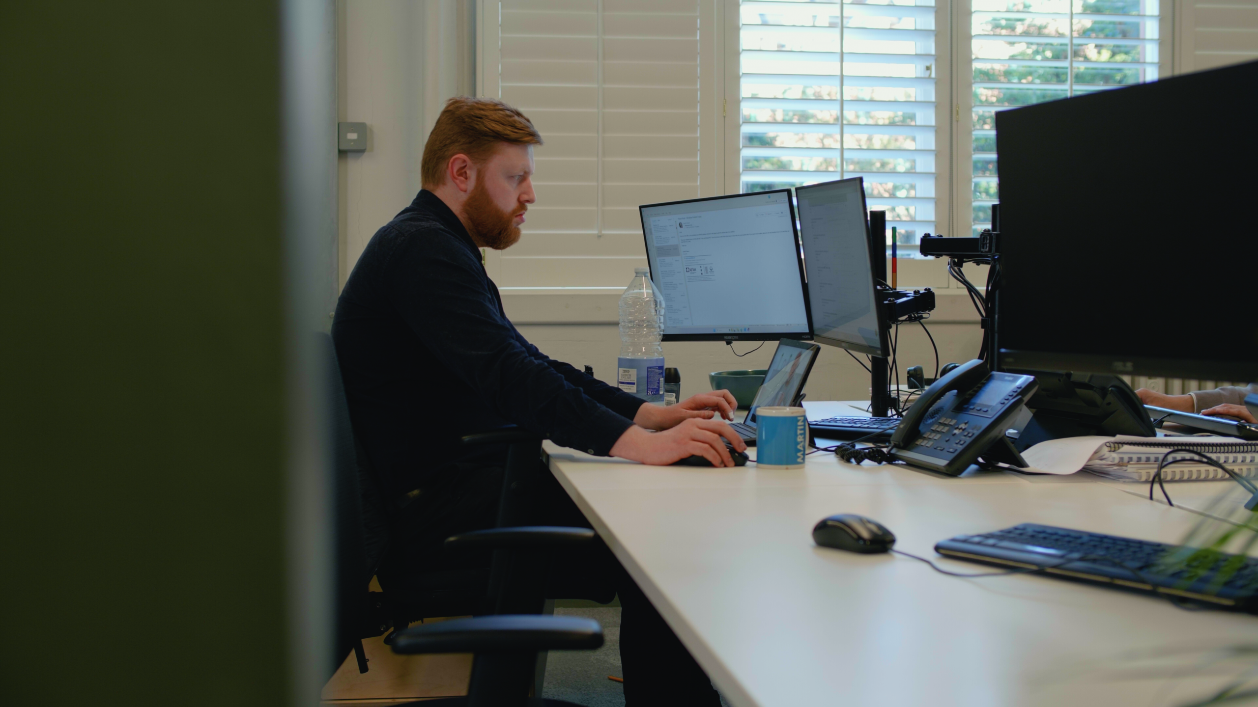 Man working at desk with dual monitors, laptop, and phone.